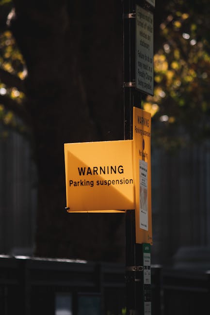 A close-up of a yellow warning sign attached to a black metal pole, indicating a parking suspension, with the text 'WARNING Parking suspension' in black letters. In the background, there is a blurred view of a wooden fence, trees with foliage, and a partially visible blue sign. The scene appears to be outdoors during daylight, with natural lighting casting soft shadows. The signage suggests local parking restrictions which may impact house removals or vehicle access during relocation activities, relevant to services offered by Man with Van Cranford. The focus is on the signage, highlighting parking suspension notices that could affect vehicle parking during home relocation or furniture transport services.