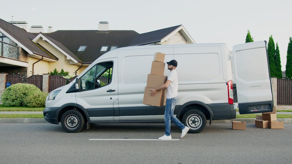 A man with a beard, wearing a black cap, white t-shirt, blue jeans, and white sneakers, is seen loading medium-sized cardboard boxes onto a white commercial van parked on a residential street. The man is holding three boxes stacked vertically, preparing for a home relocation or furniture transport within the context of packing and moving. The van has its rear doors open and additional boxes on the pavement nearby, indicating active moving or removal procedures. The setting features a row of modern, detached houses with pitched roofs, chimneys, and well-maintained gardens, with greenery and tall trees visible in the background. The scene is captured in daylight with clear weather, highlighting the loading process and equipment used during house removals. This typical residential environment exemplifies the logistics involved in professional removals services, such as those offered by Man with Van Cranford.