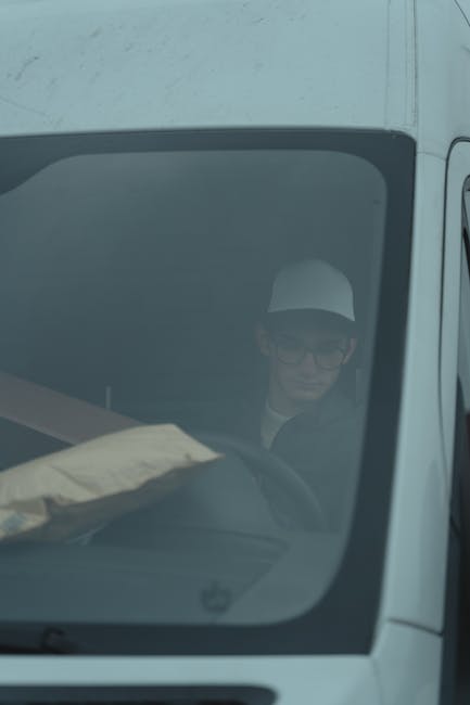 A man wearing glasses and a white cap is seated inside a white moving van, with his face partially visible through the front windshield. He appears to be engaged in his task, possibly overseeing the loading process or preparing for a home relocation. The van's interior shows a glimpse of packing materials, including a cardboard box and protective blankets, indicating the ongoing transfer of furniture and household items. The vehicle is parked on a driveway or street, and the image suggests a professional removals context carried out by Man with Van Cranford, specialised in furniture transport and packing services. The lighting is neutral, highlighting the interior and the man’s focus on the relocation process, which is part of efficient and careful moving and packing logistics.