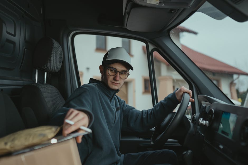A man with a beard, wearing a black cap, white t-shirt, blue jeans, and white sneakers, is seen loading medium-sized cardboard boxes onto a white commercial van parked on a residential street. The man is holding three boxes stacked vertically, preparing for a home relocation or furniture transport within the context of packing and moving. The van has its rear doors open and additional boxes on the pavement nearby, indicating active moving or removal procedures. The setting features a row of modern, detached houses with pitched roofs, chimneys, and well-maintained gardens, with greenery and tall trees visible in the background. The scene is captured in daylight with clear weather, highlighting the loading process and equipment used during house removals. This typical residential environment exemplifies the logistics involved in professional removals services, such as those offered by Man with Van Cranford.