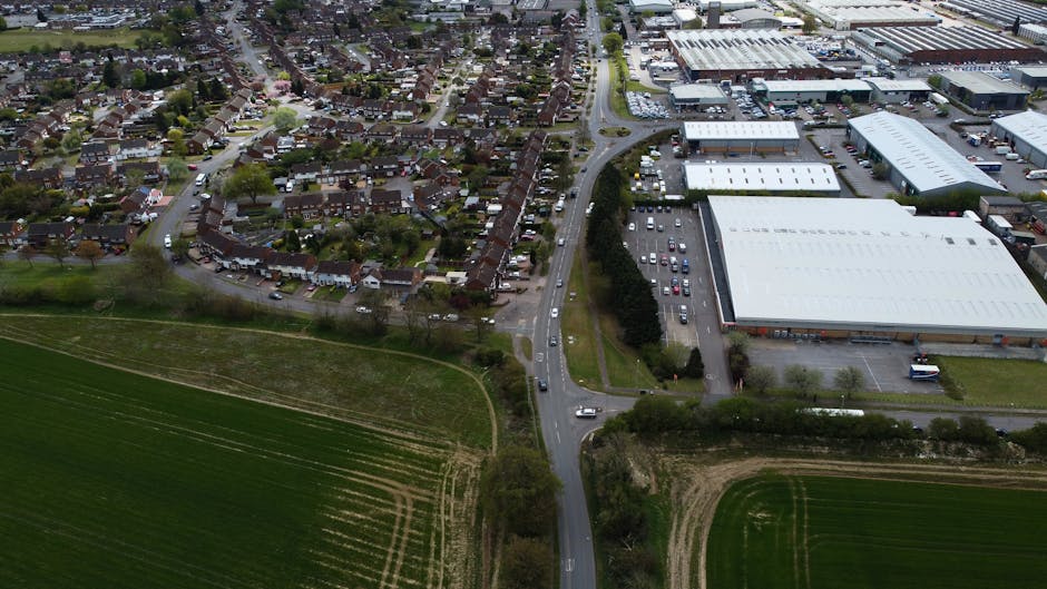 An aerial view of a residential area and commercial industrial buildings showing a curved road with several vehicles, including a white van used for home relocation or furniture transport, parked along the street and in the parking lots. The residential area features closely packed houses with gardens and trees, while the industrial zone has large warehouse-style structures with white roofs and surrounding parking areas. Green fields and open spaces border the road, providing a clear contrast between the urban and rural elements. The scene illustrates the logistics involved in house removals and moving services, with visible access points to properties and loading zones, as managed by Man with Van Cranford, supporting efficient packing and loading processes for local house moves at Cranford Lane versus Bath Road.