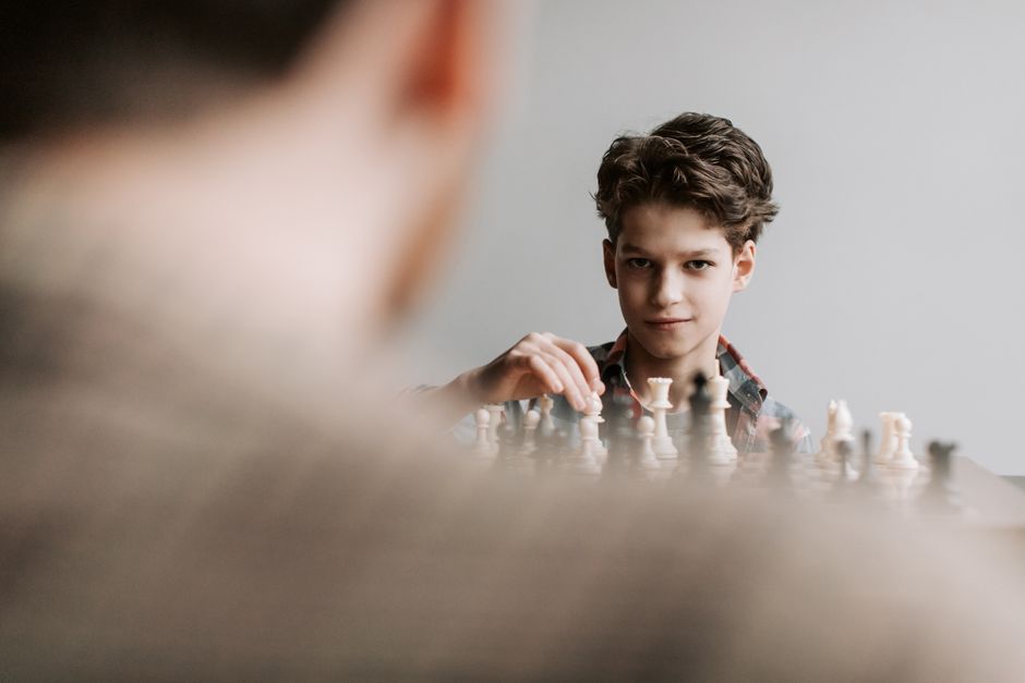 The image shows a young man with dark, wavy hair looking directly at the camera with a slight smile while playing chess on a table. In the foreground, there is a blurred figure of another person, likely an older man, with only part of their head and shoulder visible, facing the chessboard. The chess pieces are arranged on a standard wooden chessboard, and the background is a plain, neutral grey. The setting appears to be indoors with soft lighting. Although the scene depicts a leisure activity, the image can be related to the process of planning or strategic thinking involved in home relocation or moving logistics, subtly connecting to the theme of house removals and furniture transport as part of a professional moving service by Man with Van Cranford.
