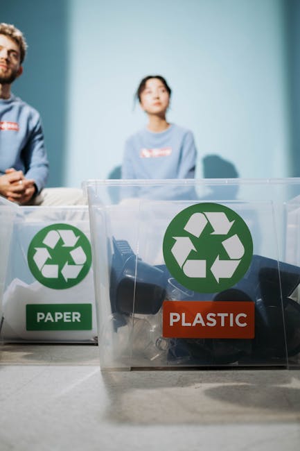 A close-up view of a clear plastic recycling bin divided into sections labeled 'PAPER' and 'PLASTIC,' with the plastic compartment containing discarded plastic objects such as bottle caps and wrappers. Behind the bins, two individuals are seated at a table in an indoor setting, with one person partially visible on the left and a woman in a light blue shirt on the right, both out of focus. The scene emphasizes eco-friendly waste disposal, which relates to environmentally conscious packing and packing material recycling involved in house removals and relocation services. The background features a plain light blue wall, and natural lighting illuminates the scene, highlighting the recycling symbols and the separation of materials, supporting the context of responsible disposal during home moves managed by Man with Van Cranford.