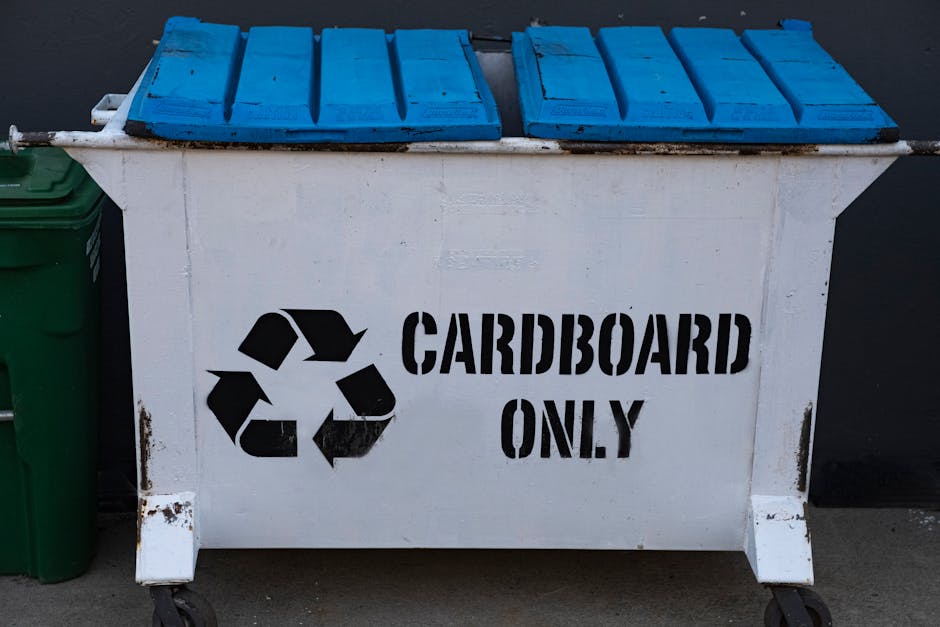 A large white wheeled waste container designated for recycling cardboard, with the recycling symbol and the words 'CARDBOARD ONLY' stenciled in black on its side. The container has a blue lid with multiple sections, positioned in an outdoor setting near a dark background, likely pavement or concrete. Next to the container, on its left side, a green bin is partially visible. The container appears slightly weathered with some rust marks near the edges, and it is situated in an area that suggests it is used for environmentally friendly disposal or recycling during a house move or relocation process. The image reflects a focus on eco-conscious packing and waste management activities, supporting efficient logistics in home removals managed by Man with Van Cranford as indicated on the website.