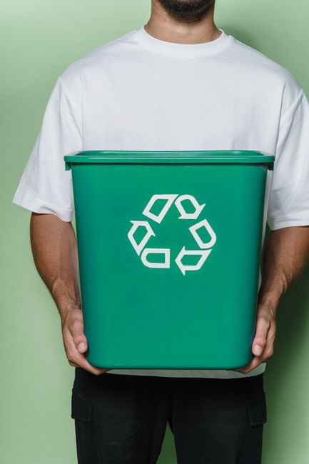 A close-up view of a clear plastic recycling bin divided into sections labeled 'PAPER' and 'PLASTIC,' with the plastic compartment containing discarded plastic objects such as bottle caps and wrappers. Behind the bins, two individuals are seated at a table in an indoor setting, with one person partially visible on the left and a woman in a light blue shirt on the right, both out of focus. The scene emphasizes eco-friendly waste disposal, which relates to environmentally conscious packing and packing material recycling involved in house removals and relocation services. The background features a plain light blue wall, and natural lighting illuminates the scene, highlighting the recycling symbols and the separation of materials, supporting the context of responsible disposal during home moves managed by Man with Van Cranford.
