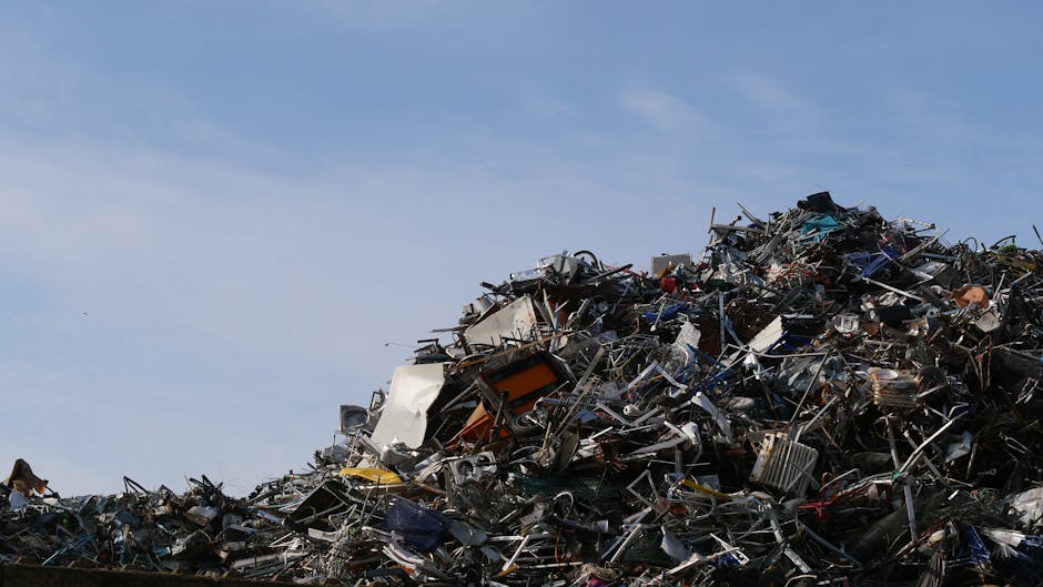 A large pile of assorted scrap metal and discarded household appliances, including metal frames, broken electronics, and damaged furniture, stacked outdoors against a clear blue sky. The debris featur