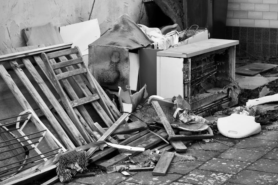 A large pile of assorted scrap metal and discarded household appliances, including metal frames, broken electronics, and damaged furniture, stacked outdoors against a clear blue sky. The debris featur
