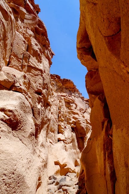 There are no visible objects related to house removals or moving services in this image. The photo depicts a narrow, rocky passageway between high sandstone cliffs under a clear blue sky, with no furniture, boxes, appliances, packaging materials, or moving equipment present. The scene appears outdoors in a natural environment, with sunlight illuminating the textured rock surfaces. As there are no signs of a home, interior space, or loading process, this image does not support content related to home relocation or furniture transport, and it does not align with moving logistics or packing activities. Therefore, no description relevant to house removals or moving services from Man with Van Cranford can be provided based on this visual.