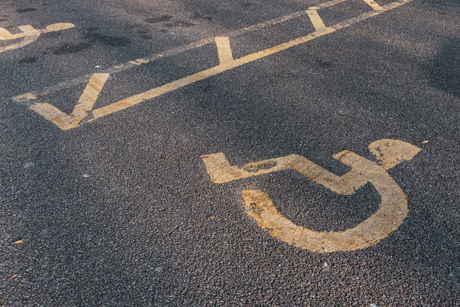A close-up view of an asphalt parking area displaying a yellow-painted wheelchair accessibility symbol and directional lines. The paint appears slightly worn and faded, indicating frequent use. The surface has a textured appearance with small stones embedded in the dark grey asphalt. There are no vehicles present in the image. This scene is relevant to house removals and moving services, illustrating parking considerations for loading and unloading in urban environments. Man with Van Cranford often manages logistics involving vehicle access and parking restrictions during home relocation or furniture transport. Properly designated disabled parking zones facilitate efficient loading and unloading processes, assisting in smooth moving operations. The image emphasizes the importance of clear signage and surface markings for vehicle positioning during furniture transport or packing and moving activities, especially in areas with limited access or narrow entryways, which may require specialized strategies during a move from Bath Road TW5.