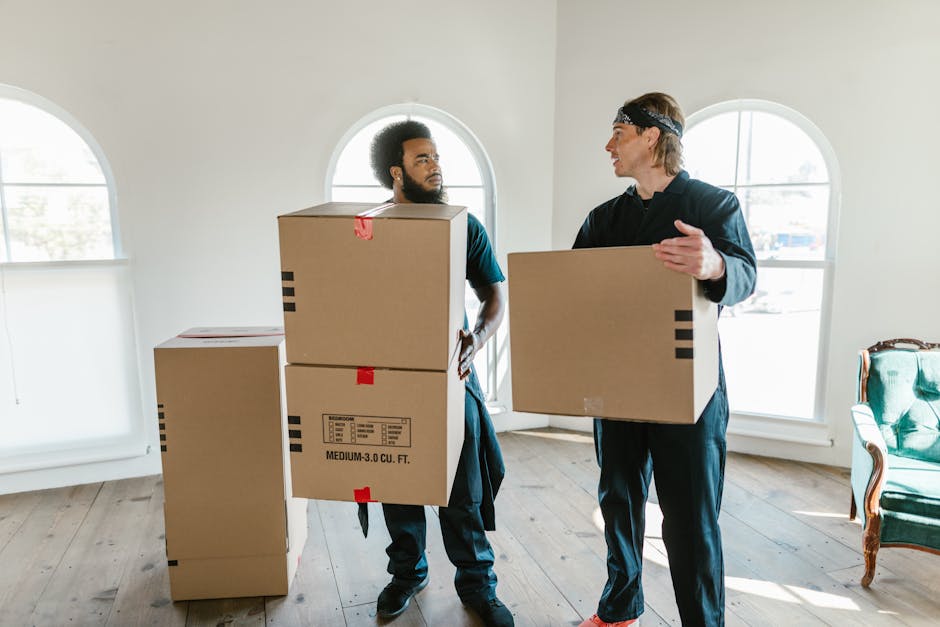 Two professional movers from Man with Van Cranford are inside a well-lit room with large arched windows, preparing for a house removal. The mover on the left, with a beard and curly hair, is holding two stacked medium-sized cardboard boxes labeled with handling and content information. The other mover, with blond hair and wearing a headband, is carrying a single cardboard box of similar size. Several additional boxes, some sealed with red tape, are placed on the wooden floor near them, indicating an active packing and loading process. The room's neutral walls and natural lighting provide a clear view of the furniture and packaging materials used during the furniture transport or home relocation. An ornate armchair with green upholstery and wooden trim is visible on the right, adding to the domestic setting. This scene illustrates the logistics of packing and moving, emphasizing the careful handling of boxes and the planning involved in home removals, supported by [COMPANY_NAME]'s professional services.