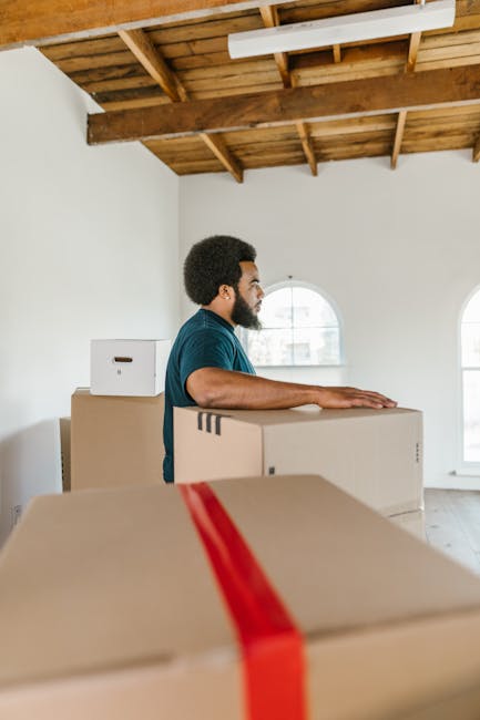 A man with a dark curly hair and beard wearing a blue T-shirt is seen in an unfinished room with a wooden ceiling structure and white walls, standing behind several large cardboard boxes stacked on top of each other. The boxes, some with visible tape, are positioned on the floor and are being prepared for a house removal or furniture transport. The room has natural light coming through arched windows, illuminating the space, which appears to be part of a home undergoing packing and moving. The man is either inspecting or preparing the boxes for loading, possibly as part of a same-day removals service offered by Man with Van Cranford, specializing in efficient and professional house relocation and packing and moving logistics.