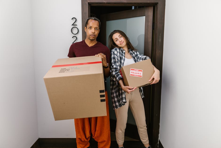Two professional movers from Man with Van Cranford are inside a well-lit room with large arched windows, preparing for a house removal. The mover on the left, with a beard and curly hair, is holding two stacked medium-sized cardboard boxes labeled with handling and content information. The other mover, with blond hair and wearing a headband, is carrying a single cardboard box of similar size. Several additional boxes, some sealed with red tape, are placed on the wooden floor near them, indicating an active packing and loading process. The room's neutral walls and natural lighting provide a clear view of the furniture and packaging materials used during the furniture transport or home relocation. An ornate armchair with green upholstery and wooden trim is visible on the right, adding to the domestic setting. This scene illustrates the logistics of packing and moving, emphasizing the careful handling of boxes and the planning involved in home removals, supported by [COMPANY_NAME]'s professional services.