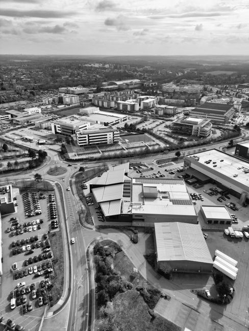 A black and white aerial photograph showing a large commercial and industrial complex with multiple low-rise buildings, some with flat roofs, surrounded by parking areas filled with numerous parked cars. The complex includes structures with varied roofing materials, some with skylights or vents, and appears to be situated near a busy roundabout connected to roads that lead to residential and urban areas in the distance. The area is well-organized with clearly marked parking bays and access roads. Trees and greenery are visible along the roads and around the edges of the complex. The scene captures the broad layout typical of a site involved in home relocation and furniture transport logistics, aligning with a professional removals service, as seen by the nearby roads and infrastructure supporting moving activities, consistent with the content on manwithvancranford.co.uk.