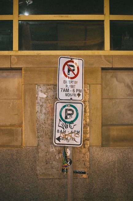 Close-up of a metal parking restriction and permit sign mounted on a wall and supported by a pole, with a beige stone wall and a large window with wooden framing in the background. The top sign displays a no-parking symbol with text indicating parking is restricted except by permit from 7 am to 6 pm, Monday to Friday, with a black arrow pointing right. The lower sign features a 'P' symbol encircled in blue, with additional identification markings and a black arrow showing parking is allowed from 8 am to 6 pm, Monday to Friday. The signs are attached to a pole that has some labels and a strap hanging from it. The setting appears to be outside a building, possibly a commercial or residential property, with the signs indicating parking regulations relevant for a house removal or relocation project managed by Man with Van Cranford, as part of the logistical planning for furniture transport and loading during a house removal process.