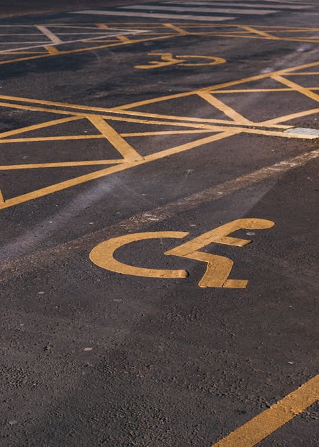 A close-up view of a paved parking area showing yellow painted wheelchair accessible parking symbols on the asphalt surface, with multiple diagonal and horizontal lines indicating parking bays and loading zones. The surface appears clean and well-marked, consistent with designated parking spaces used during house removals or furniture transport. The scene is outdoors, with natural lighting illuminating the markings. This image illustrates the importance of parking permits and designated loading areas for efficient and compliant home relocation services, as provided by Man with Van Cranford. The markings help facilitate the loading process in residential or commercial settings, supporting smooth logistics during house removals or moving operations.